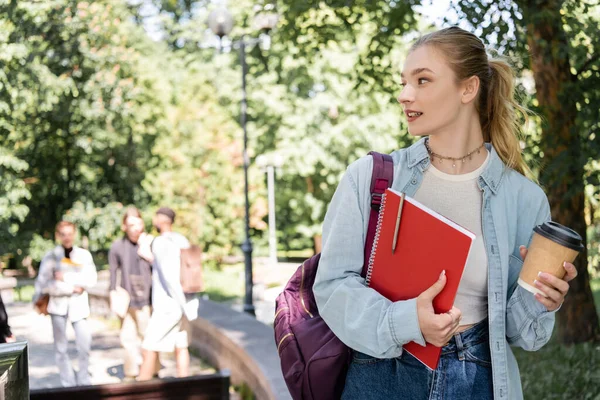 Joven estudiante con bebida para llevar y cuaderno de pie en el parque borroso - foto de stock