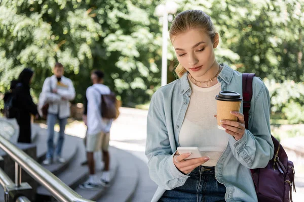 Young student with backpack using smartphone and holding takeaway drink in park - foto de stock