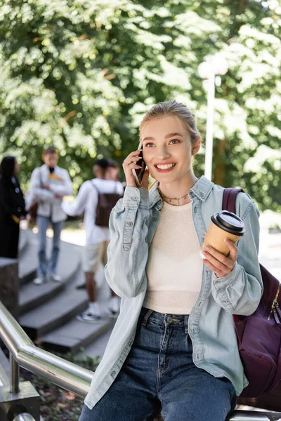 Estudante sorrindo com café para ir e mochila falando no smartphone no parque — Fotografia de Stock