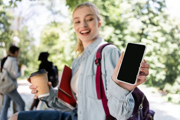 Estudante desfocado com café para ir mostrando smartphone com tela em branco no parque — Fotografia de Stock