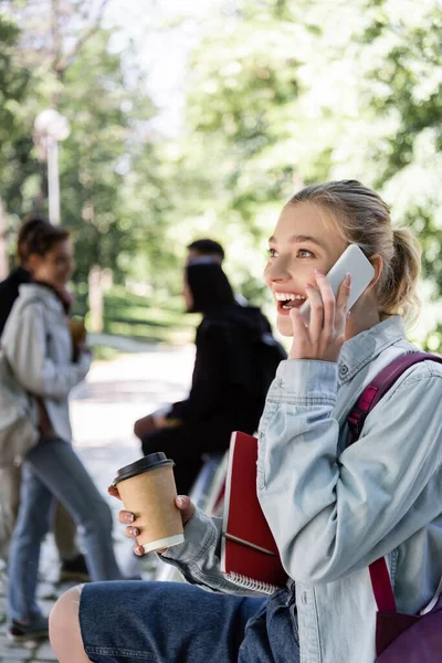 Excited student with coffee and notebook talking on smartphone in park — Stock Photo