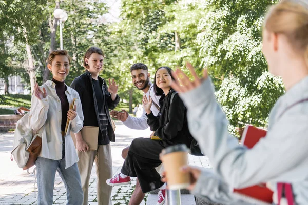 Smiling multiethnic students waving hands at blurred friend in park — Stockfoto