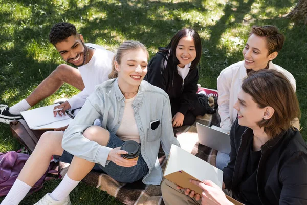 Vista de ángulo alto de estudiantes multiculturales alegres con libros de texto y dispositivos sentados en la hierba en el parque - foto de stock