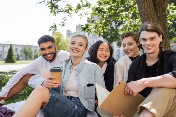 Positive multikulturelle Studenten mit Notizbuch und Kaffee blicken auf Gras im Park in die Kamera — Stockfoto