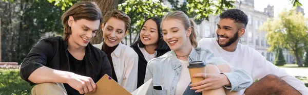 Étudiants multiethniques positifs regardant un ami avec le livre de copie dans le parc, bannière — Photo de stock