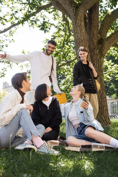 Positive interracial students with backpacks looking at friends on blanket in park — Stockfoto