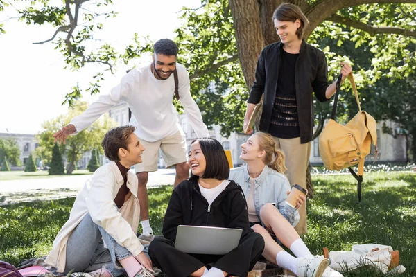 Багатоетнічні студенти з ноутбуком сидять на ковдрі біля друзів у парку — стокове фото