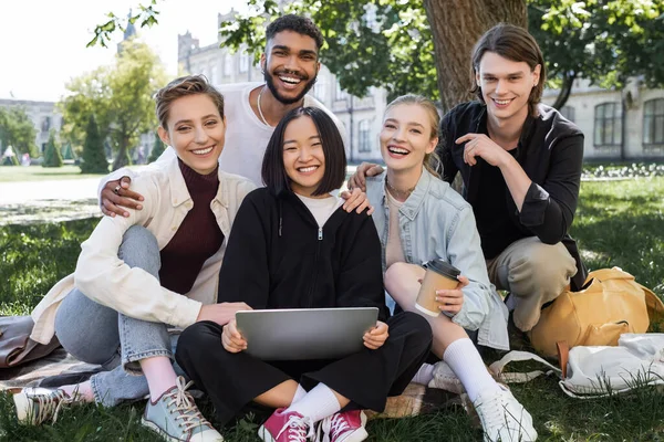 Positive multicultural students with laptop hugging on grass in park — Stockfoto