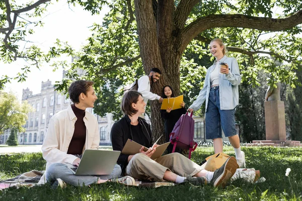 Studente con caffè per andare e zaino parlando con gli amici interrazziale nel parco estivo — Foto stock