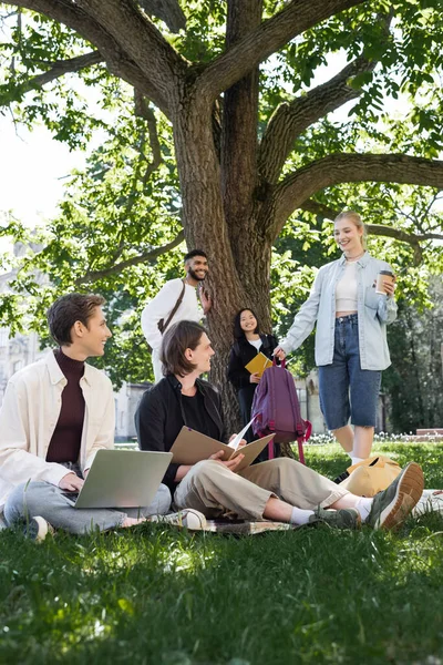 Cheerful student with takeaway drink standing near multiethnic friends on grass in summer park — Photo de stock