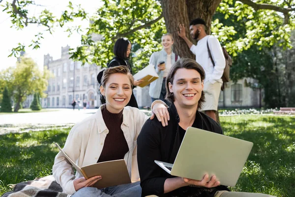 Smiling students with notebook and laptop looking at camera on grass in park — Stock Photo