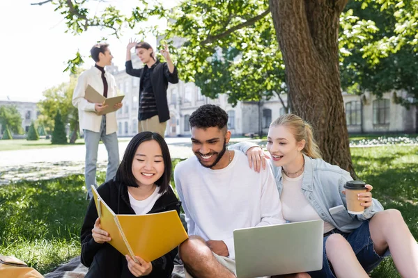 Asian student holding notebook near friends wit laptop and coffee in park - foto de stock