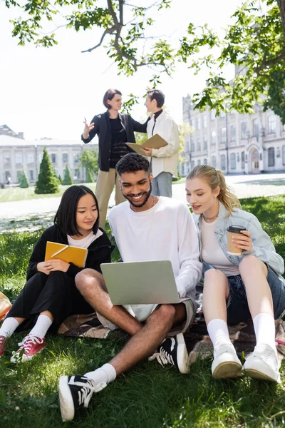 Estudiante afroamericano usando portátil cerca de amigos multiétnicos con libro de copias y café sobre hierba en el parque - foto de stock
