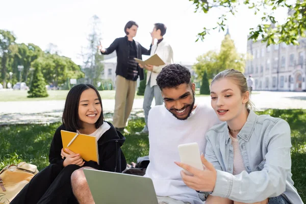 Student holding smartphone near smiling multicultural students with laptop and notebook on grass in park - foto de stock