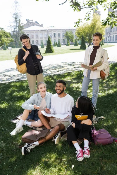 Alegre interracial estudiantes con libros de texto pasar tiempo en la hierba en el parque - foto de stock