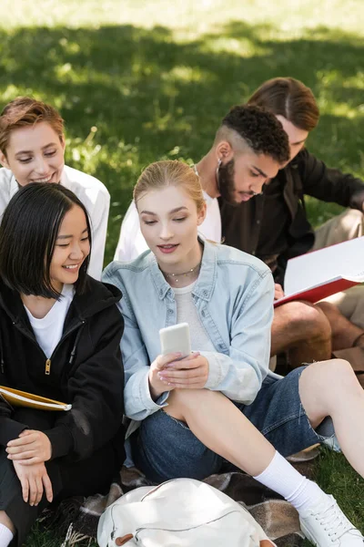 Estudiante sosteniendo teléfono inteligente cerca de amigos multiculturales en el césped en el parque de verano - foto de stock