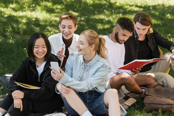 Estudiante positivo sosteniendo teléfono inteligente cerca de amigos multiétnicos en la hierba al aire libre - foto de stock