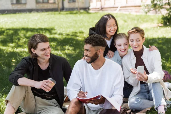 Улыбающиеся межрасовые студенты разговаривают рядом с размытыми друзьями, используя смартфон на газоне в парке — стоковое фото