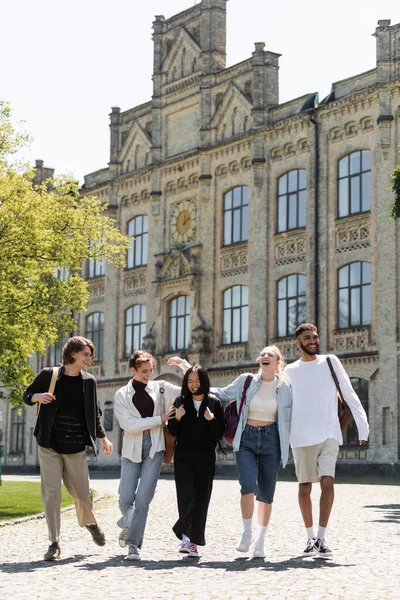Positive multiethnic students with backpacks walking near blurred university outdoors — Photo de stock
