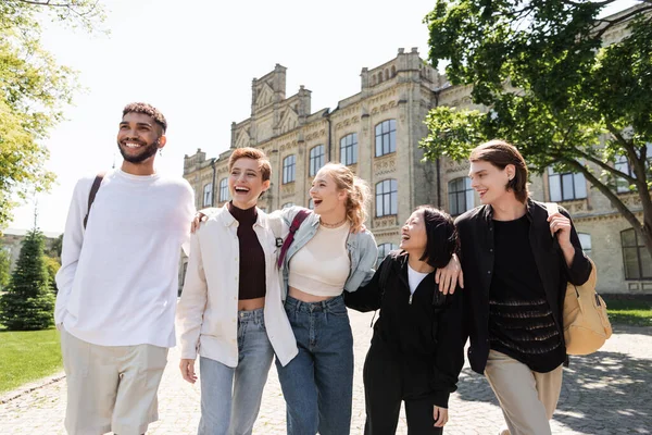 Grupo de estudantes multiculturais sorridentes abraçando enquanto caminham perto da universidade ao ar livre — Fotografia de Stock