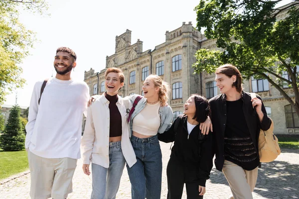 Group of smiling multicultural students hugging while walking near university outdoors — Fotografia de Stock