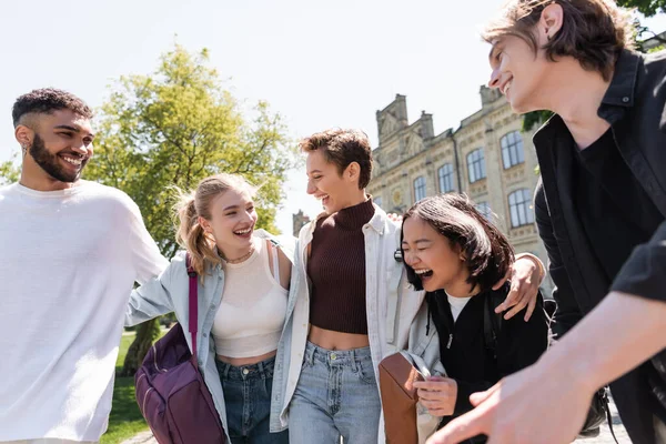 Estudante sorrindo abraçando amigos inter-raciais com mochilas ao ar livre — Fotografia de Stock