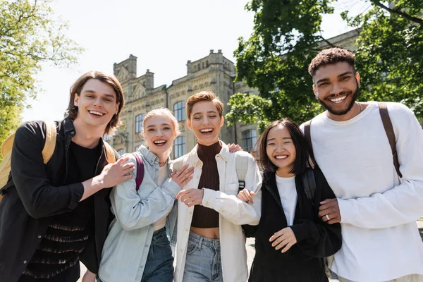 Estudiantes multiétnicos positivos abrazando y mirando a la cámara al aire libre - foto de stock