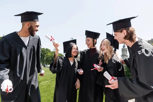 Happy interracial bachelors in caps holding diplomas in park — Stock Photo