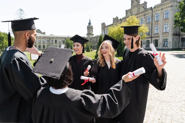 Positive multiethnic bachelors in caps and gowns holding diplomas in park — Foto stock