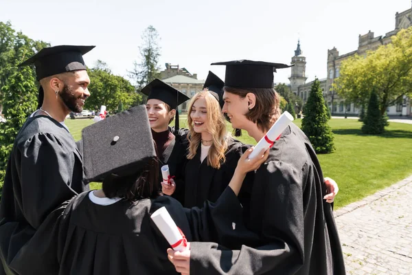 Усміхнені міжрасові холостяки в сукнях тримають дипломи і обіймаються в парку — стокове фото