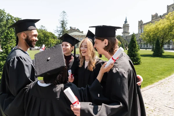 Smiling interracial bachelors in gowns holding diplomas and hugging in park — Stock Photo