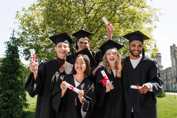 Smiling multicultural bachelors holding diplomas and looking at camera in park - foto de stock