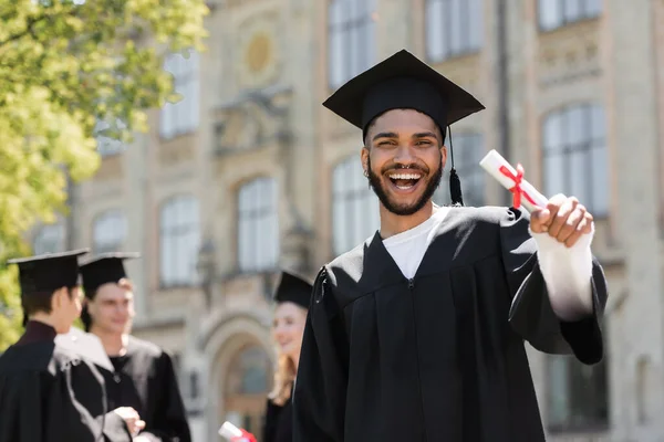 Happy african american bachelor in gown holding diploma outdoors — Stock Photo