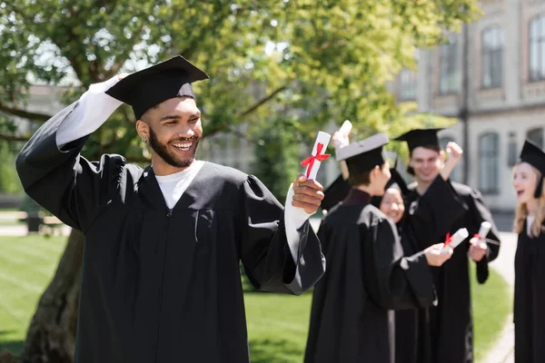 Cheerful african american bachelor looking at diploma near blurred friends in park - foto de stock