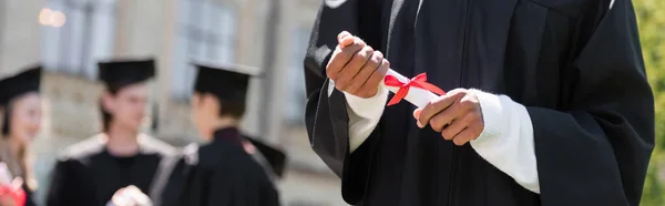 Vista ritagliata di scapolo afroamericano con diploma vicino ad amici sfocati nel parco, striscione — Foto stock