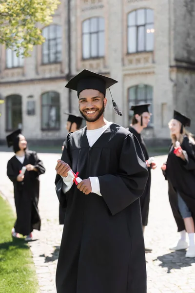 Young african american bachelor in gown holding diploma and looking at camera in park — Stockfoto