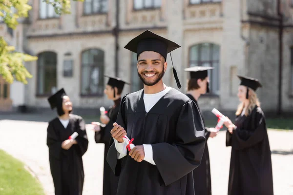 Afroamerikanischer Junggeselle in Mütze und Kleid mit Diplom in der Nähe verschwommener Freunde und Universität im Freien — Stockfoto