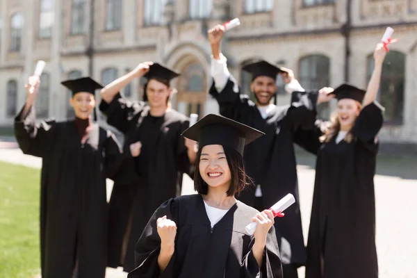 Emocionado asiático soltero celebración diploma cerca borrosa amigos en parque - foto de stock