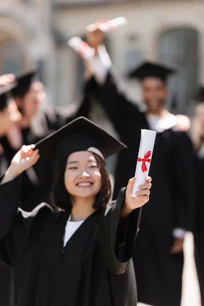 Blurred asian bachelor in gown holding diploma outdoors - foto de stock
