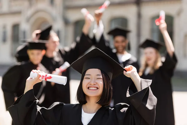 Smiling Asian bachelor in cap and gown holding diploma near blurred friends outdoors — Stockfoto