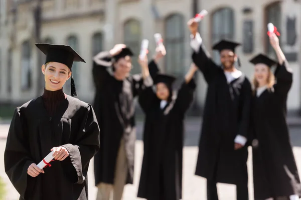 Smiling bachelor in cap holding diploma near blurred interracial friends outdoors — Foto stock