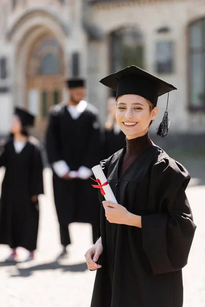 Souriant célibataire en robe détenant un diplôme et regardant la caméra à l'extérieur — Photo de stock