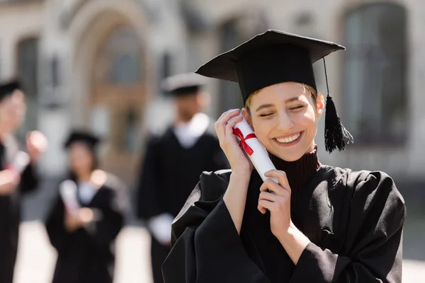Cheerful bachelor in cap holding diploma near face outdoors — Stock Photo