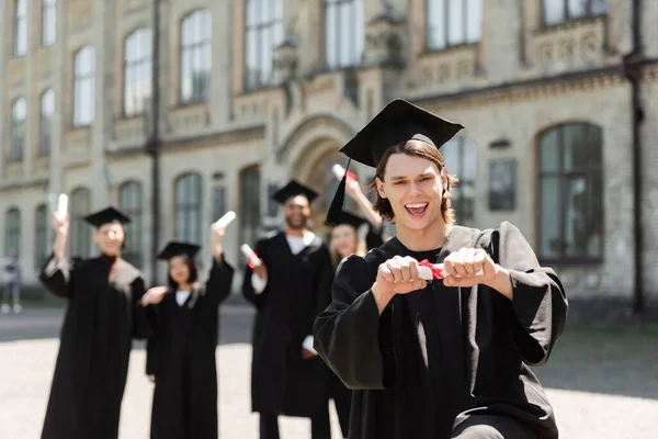 Positive bachelor holding diploma and looking at camera near blurred interracial friends outdoors — Fotografia de Stock