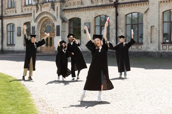 Возбужденный холостяк с дипломом, показывающим да жест рядом с межрасовыми друзьями и университетом на открытом воздухе — стоковое фото