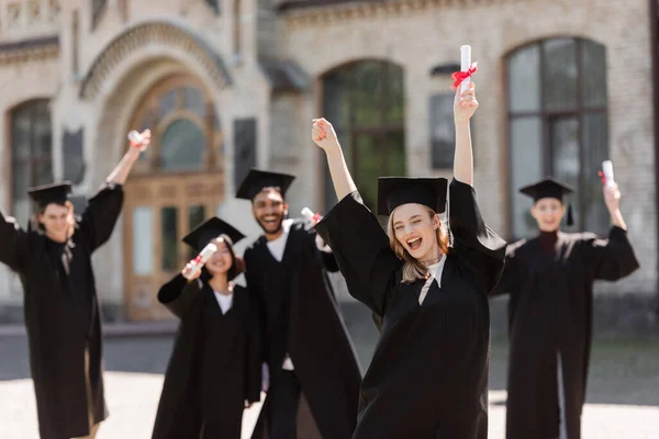 Aufgeregte Studentin im Bachelorkleid mit Diplom bei multiethnischen Freunden im Freien — Stockfoto