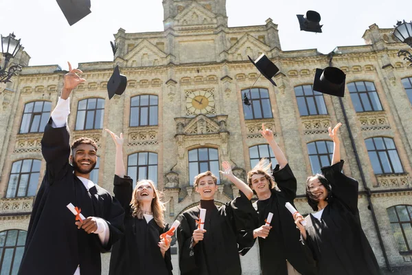 Low angle view of interracial bachelors throwing caps near university outdoors — Foto stock