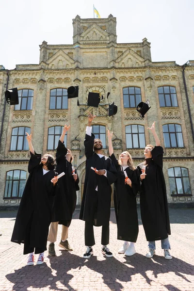 Positive interracial bachelors with diplomas throwing caps near university outdoors - foto de stock