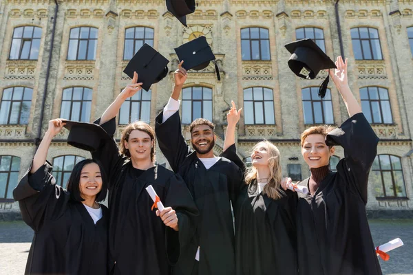 Smiling multiethnic bachelors holding diplomas and throwing caps outdoors — Stock Photo