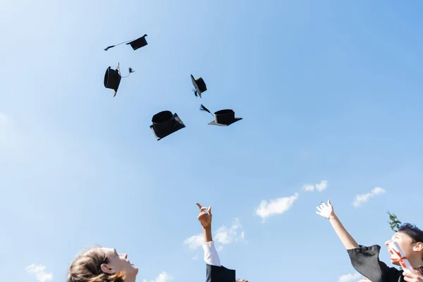 Vista de ángulo bajo de solteros multiétnicos lanzando gorras al aire libre - foto de stock